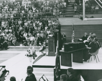 Thumbnail for 'A distinguished guest speaker addresses candidates and guests at a mid-1960s Commencement exercise in Mountaineer Gymnasium.'